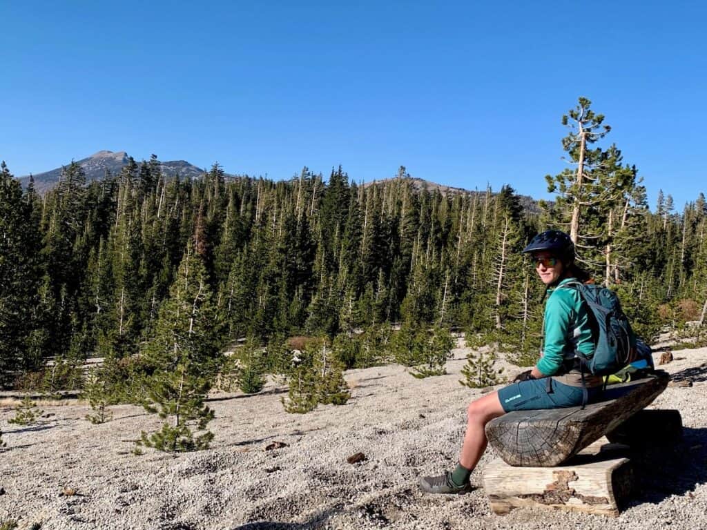 Woman sitting on bench wearing bike helmet in scenic clearing surrounded by fir trees