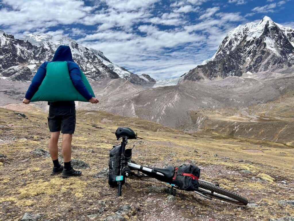 Man standing at viewpoint next to bike looking out over snow-capped mountains. Wind is billowing his jacket