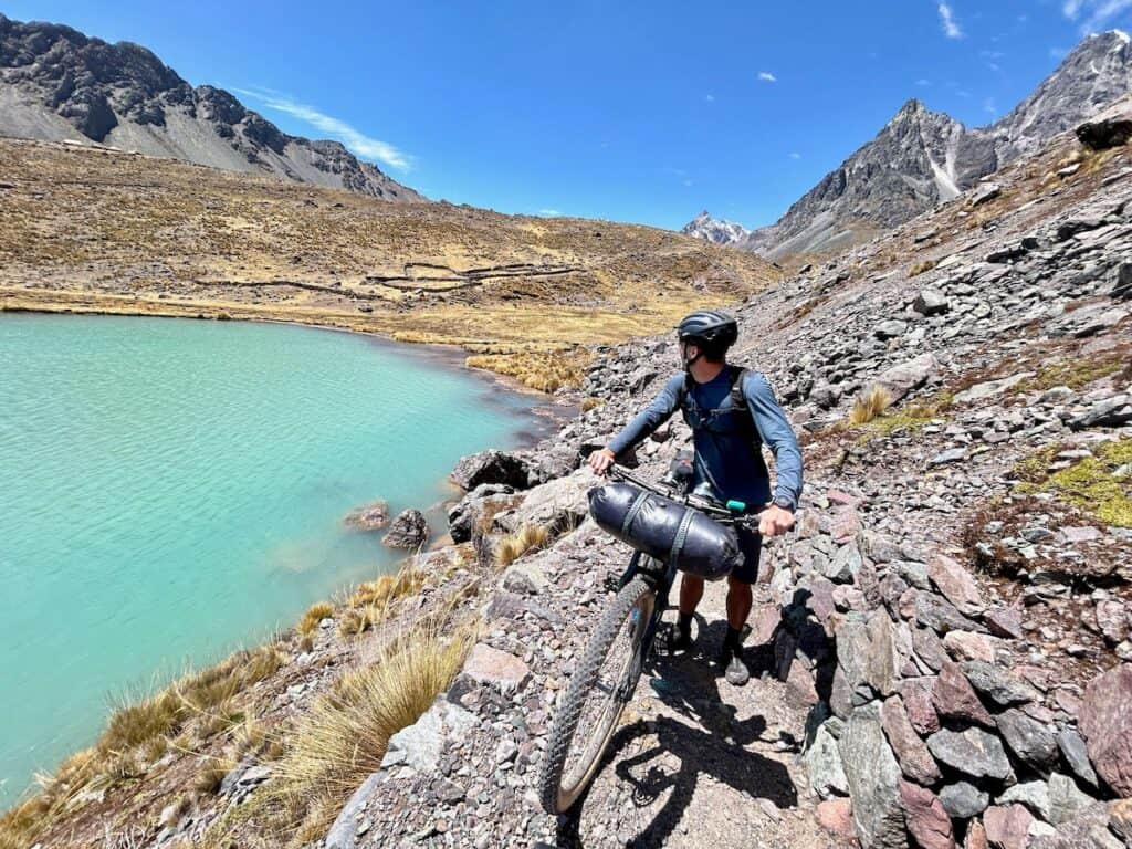 Man pushing bike up a trail looking back at turquoise colored alpine lake on the Ausangate Circuit in Peru