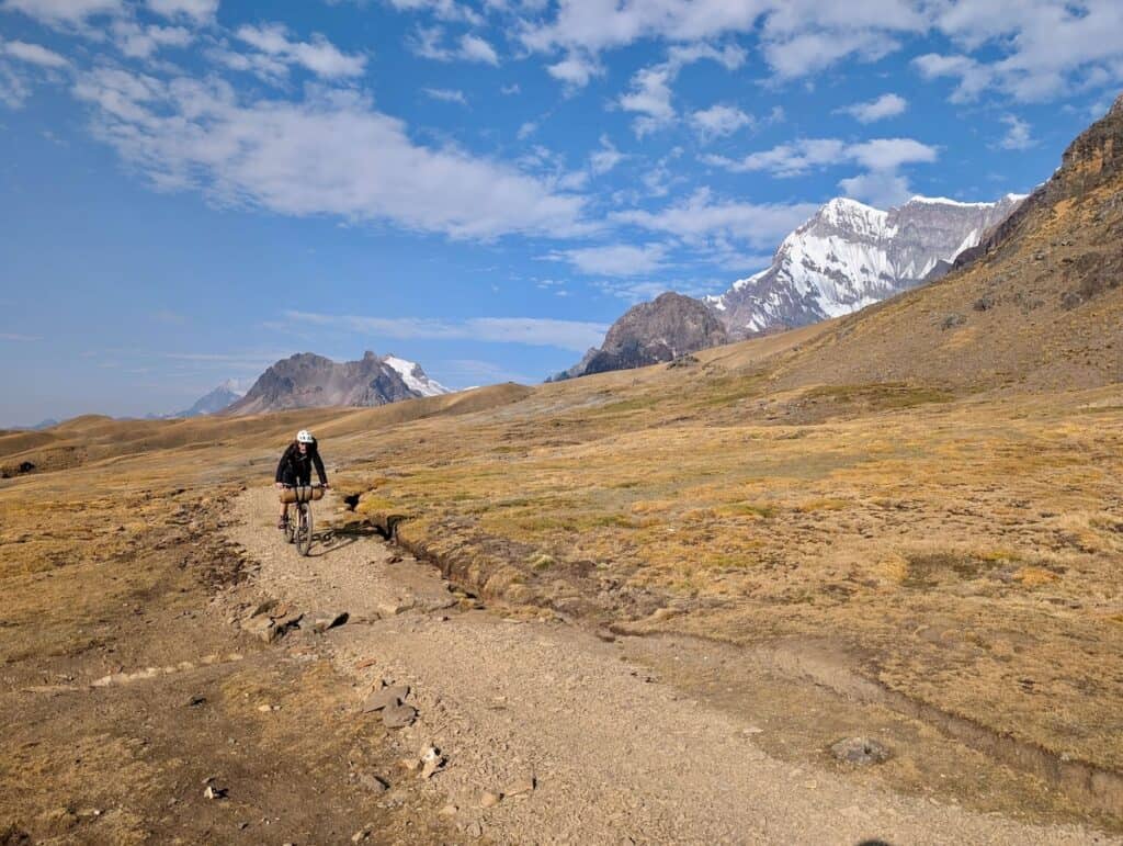 Bikepacker riding bike down wide dirt track with snow-capped mountains in the background