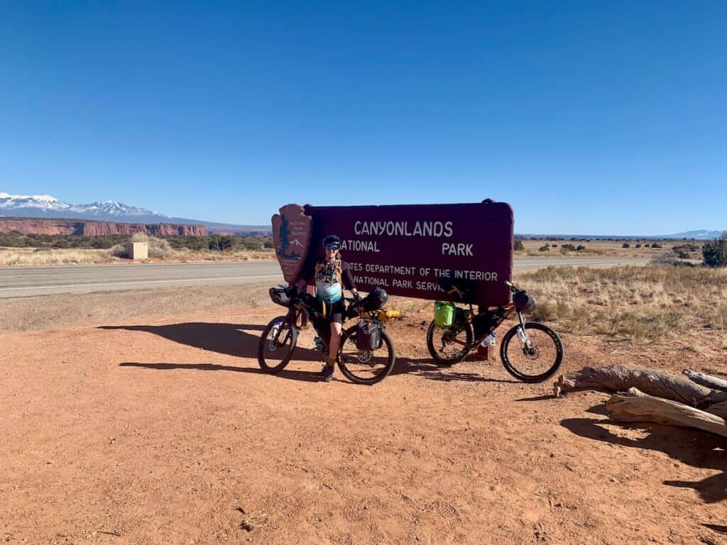 Woman standing with loaded bikepacking bike in front of Canyonlands National Park sign