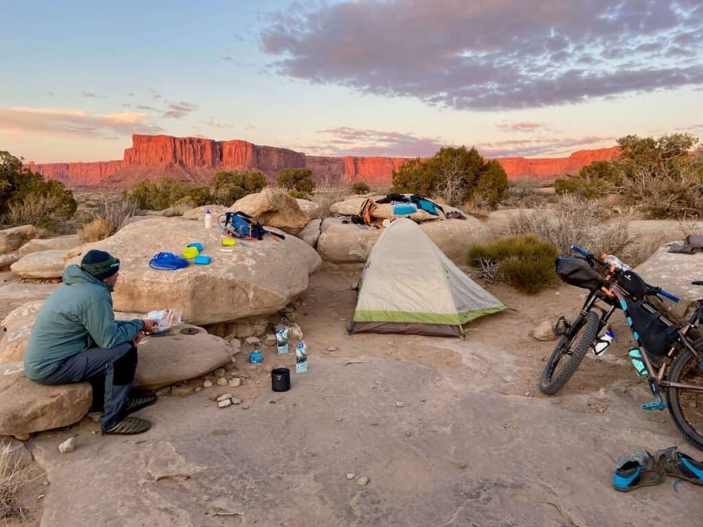 Man sitting at remote campground with gear laid out. Sun is setting on red rock bluffs and canyonlands landscape around him