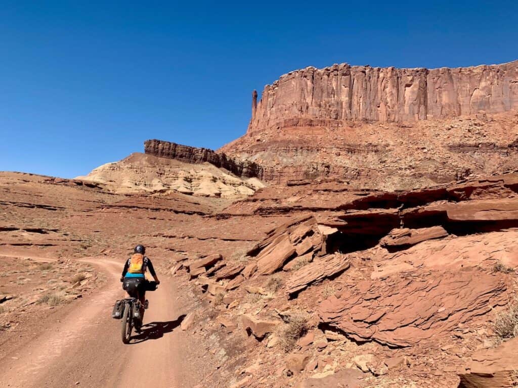 Woman riding loaded bikepacking bike up red dirt road surrounded by tall red sandstone bluffs in Utah