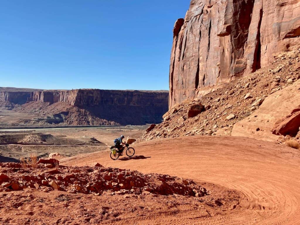 Bikepacking pedaling loaded bike up steep switchbacked road surrounded by tall red sandstone bluffs in Utah