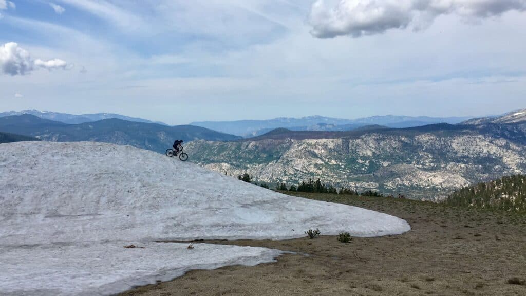 Mountain biker riding down large snow drift with beautiful mountain range views in the background