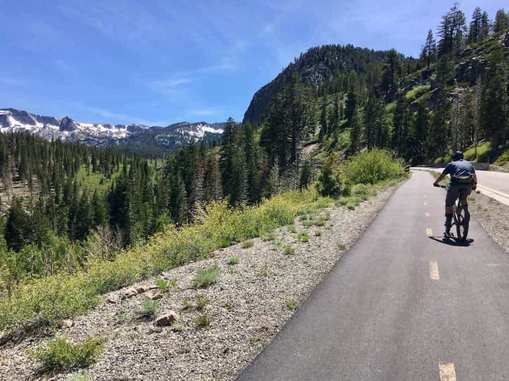 Cyclist on paved bike path lined with green trees and surrounded by beautiful mountains