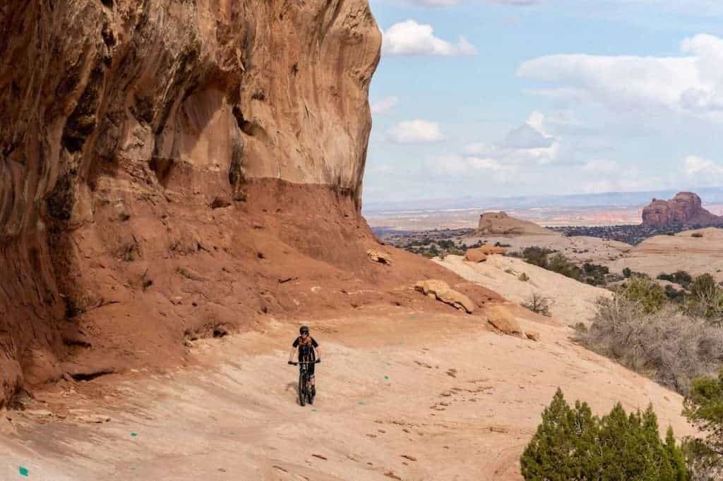 Becky riding mountain bike on slickrock section of trail in Moab, Utah with tall red rock bluff on one side