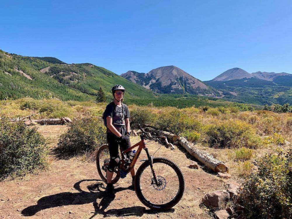 Becky posing for photo on mountain bike on The Whole Enchilada trail in Moab with green meadow and mountains behind her