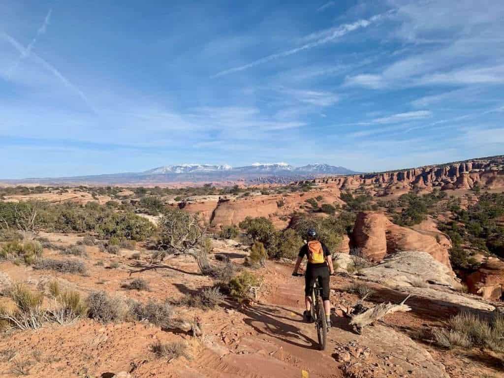 Mountain bike riding away from camera on singletrack trail in Moab, Utah surrounded by red rock formations and snow-capped mountains in the distance