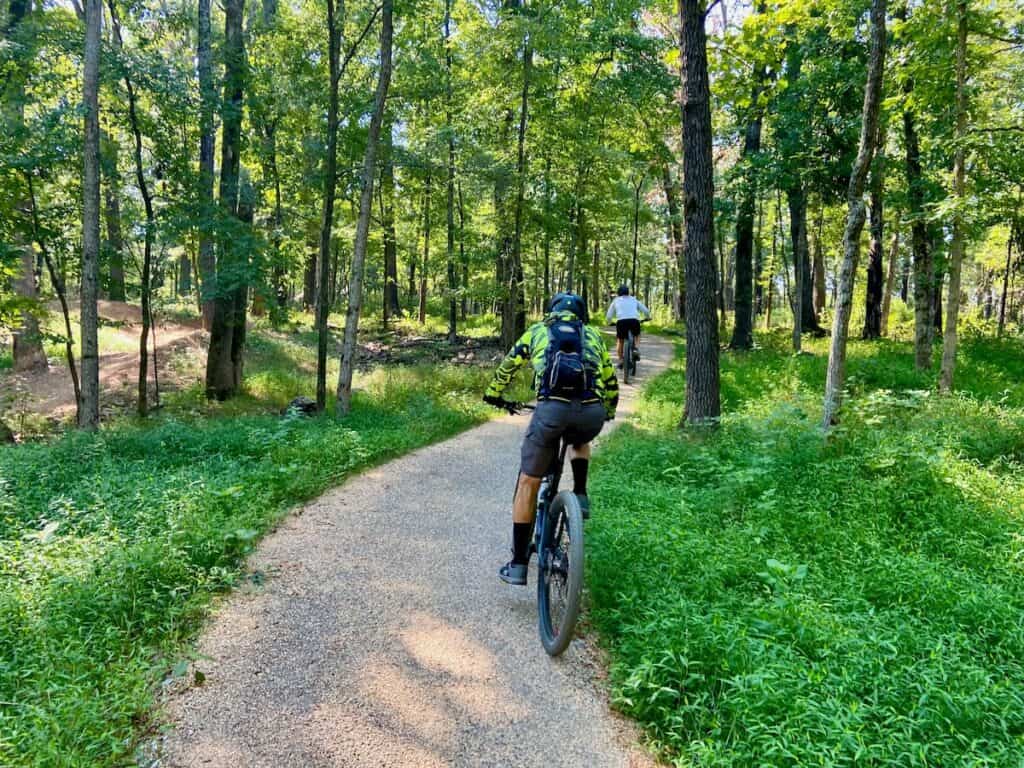 Mountain bikers pedaling up a paved path set in a lush green forest. 