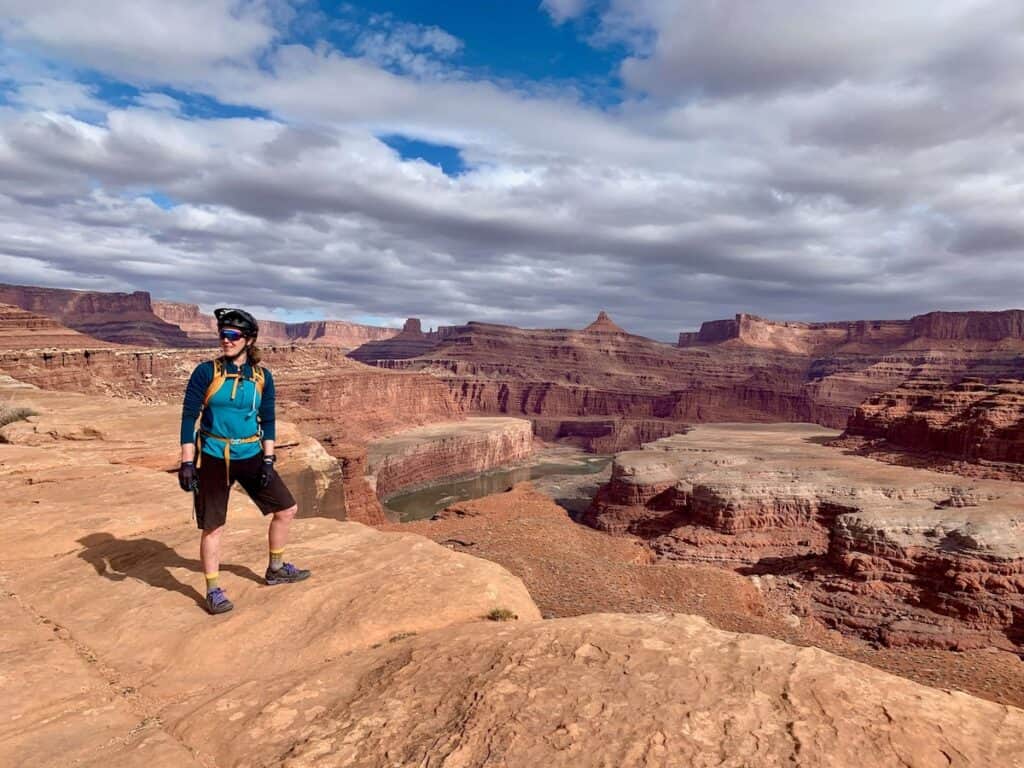 Woman wearing bicycle helmet standing at edge of stunning red canyonlands lookout on the White Rim Trail near Moab, Utah